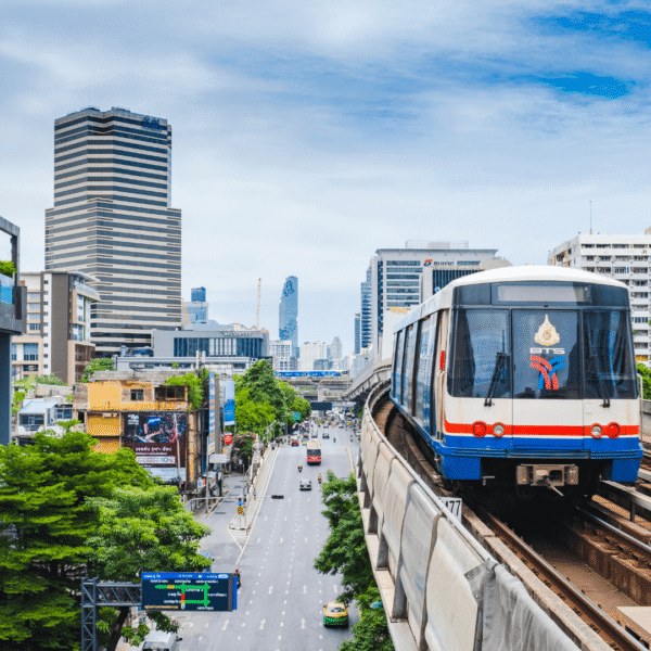 Bangkok skytrain moving on elevated tracks with city skyline, featuring modern buildings and urban traffic below, under a cloudy sky.