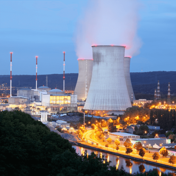 Nuclear power plant at dusk, with cooling towers emitting steam. Illuminated roads and trees in the foreground enhance the industrial landscape.